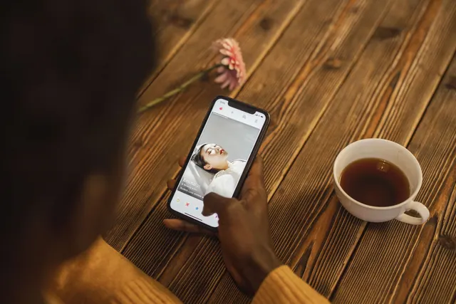 An individual using a dating app on a smartphone with a cup of tea nearby on a wooden table. The arithmetic of a viral trend hasn't changed much heading into 2027